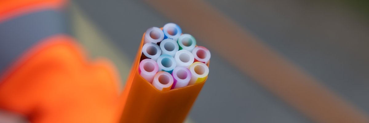 Nahaufnahme eines gelben Mikrorohrbündels mit mehreren farbigen Einzelröhrchen, das für die Verlegung von Glasfaserkabeln im Tiefbau verwendet wird. Im Hintergrund ist unscharf ein Mensch in orangefarbener Warnschutzkleidung zu erkennen.